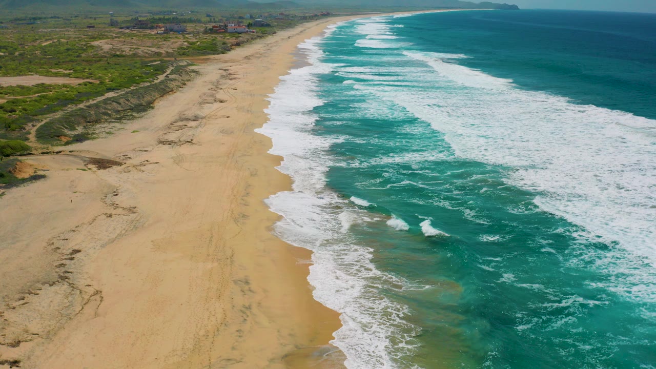 Aerial view over Cerritos, Baja California Sur, with turquoise surf reaching a broad sandy beach, scrubland inland, distant mountains, and no visible people.