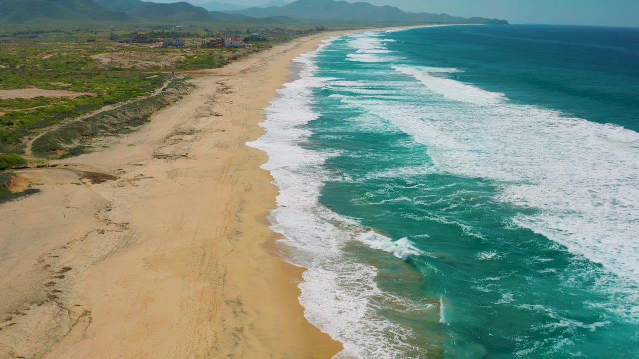 High aerial frame of the Cerritos coastline, with the beach curving into the distance, surf filling the right side, inland scrub and mountains on the left, and no visible people.