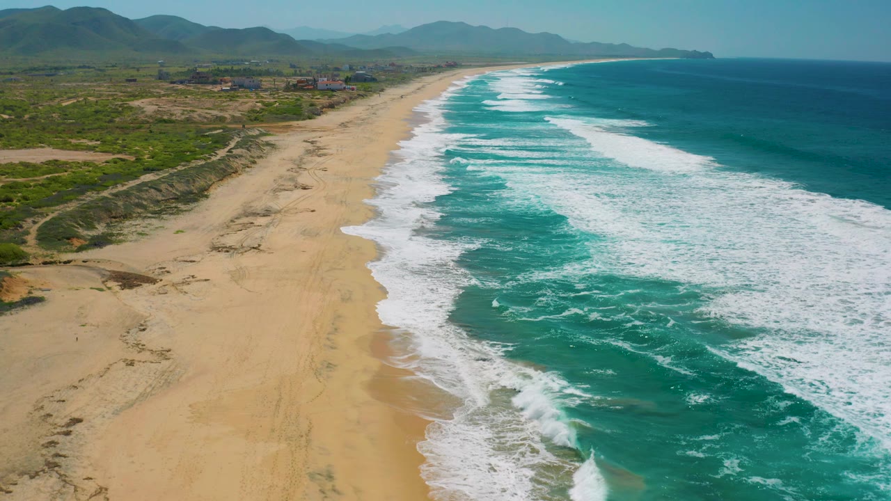Aerial view along Cerritos beach where foamy surf meets empty tan sand, with green inland terrain, distant buildings, mountain ridges, and no visible people.