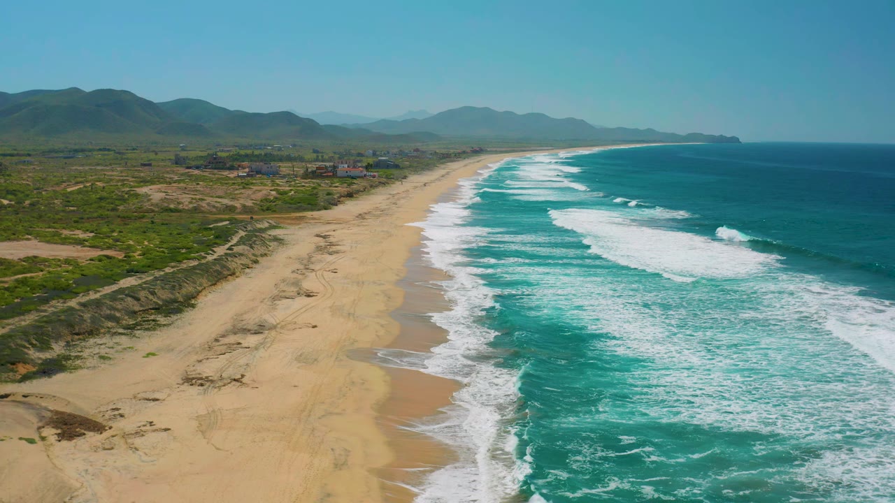 Aerial view of Cerritos coastline with white waves advancing along the beach, scrubby dunes in the foreground, scattered inland buildings, mountains, and no visible people.