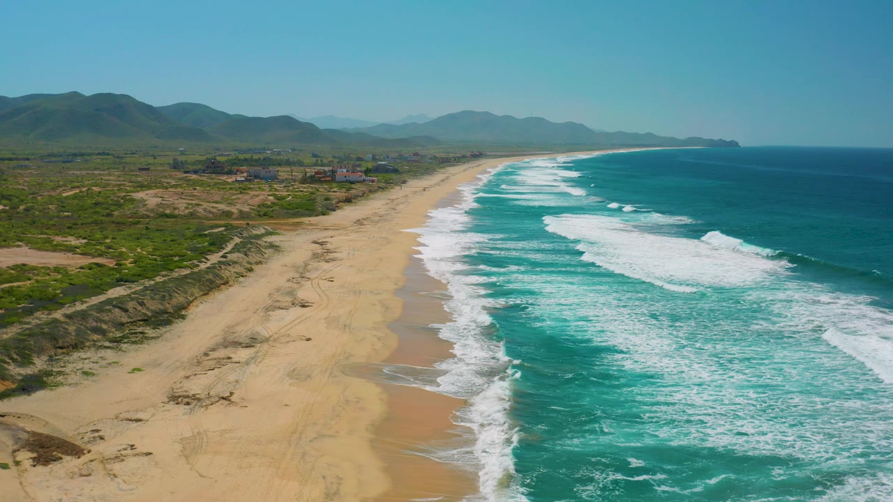 Aerial frame over Cerritos, Baja California Sur with the shoreline curving into the distance, foamy turquoise water on the right, inland greenery and mountains on the left, and no visible people.
