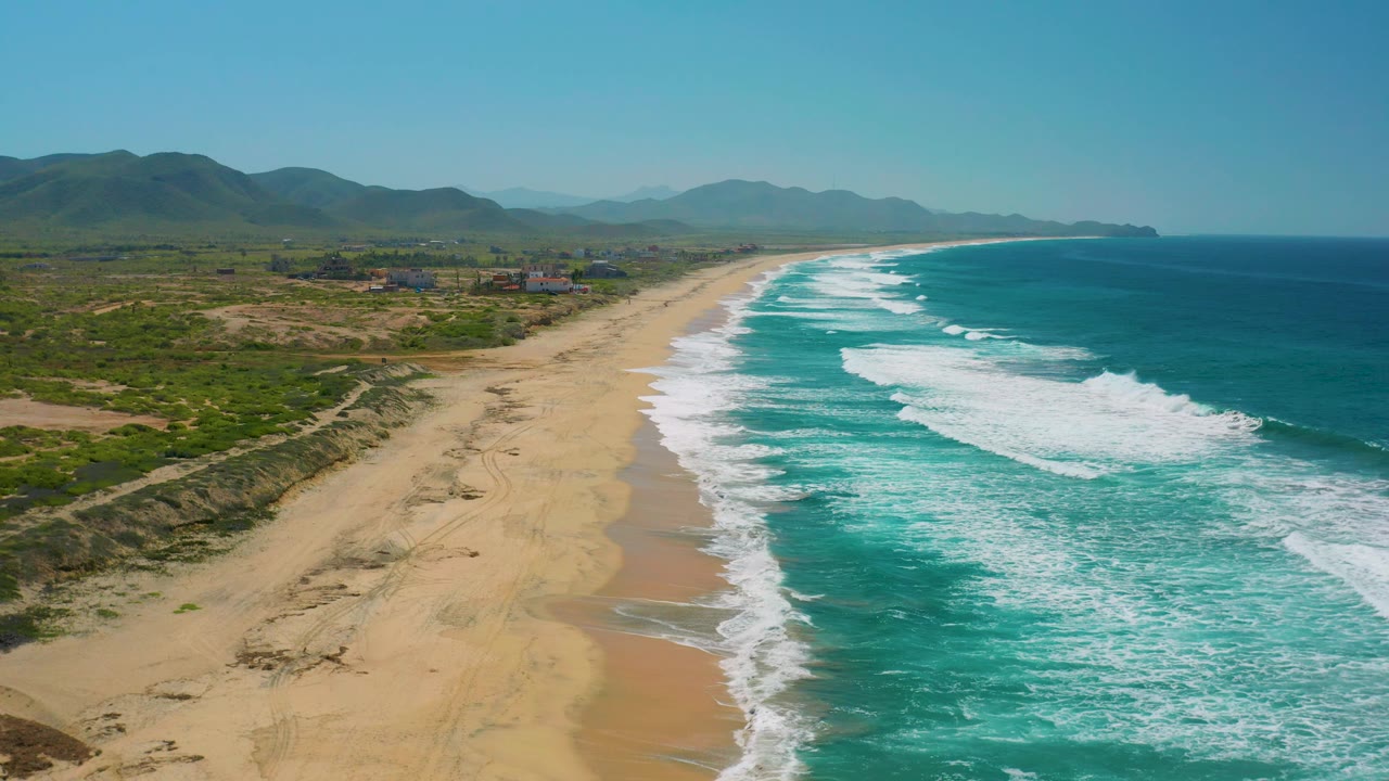 Elevated Cerritos beach frame showing dunes and scrub in the lower left, a long pale sand strip, white surf moving across turquoise water, inland buildings, mountain ridges, and no visible people.