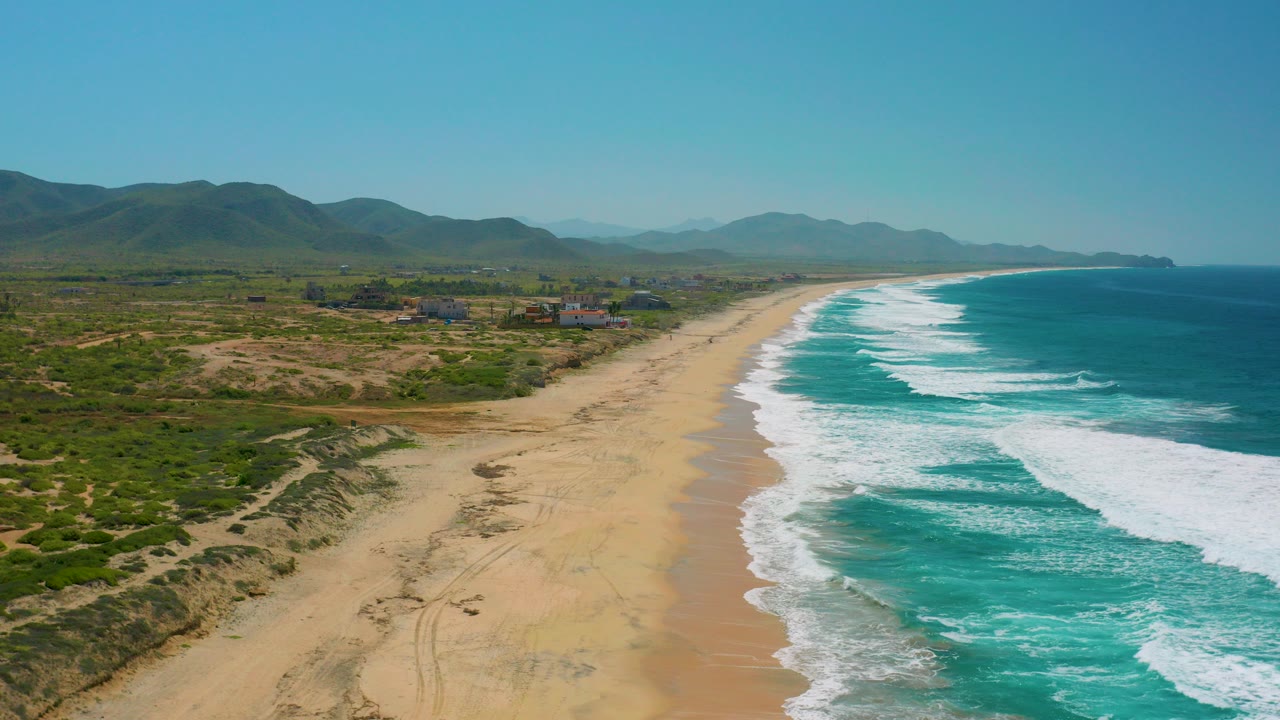 Aerial frame over Cerritos, Baja California Sur showing a long sandy beach beside turquoise surf, scrubby dunes inland, mountains on the horizon, and no visible people.
