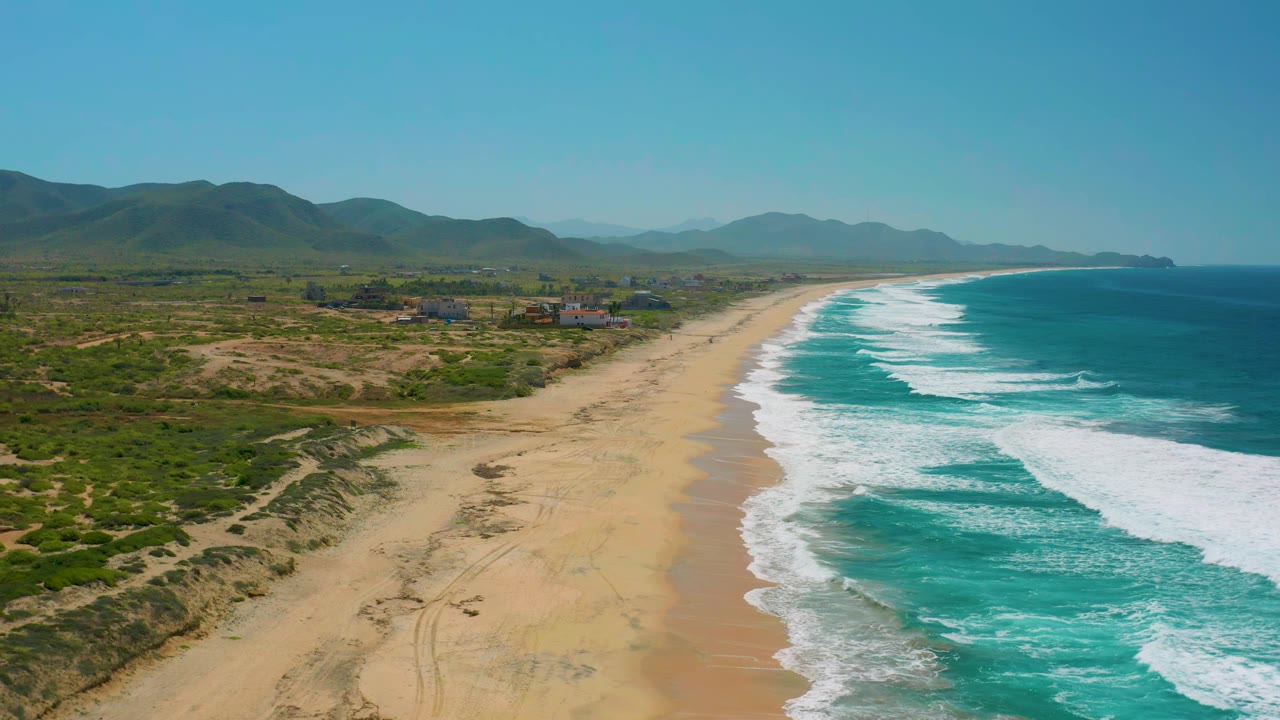 Aerial view of Cerritos beach with white waves breaking diagonally along the shore, inland scrub and sandy tracks at left, distant mountains, and no visible people.