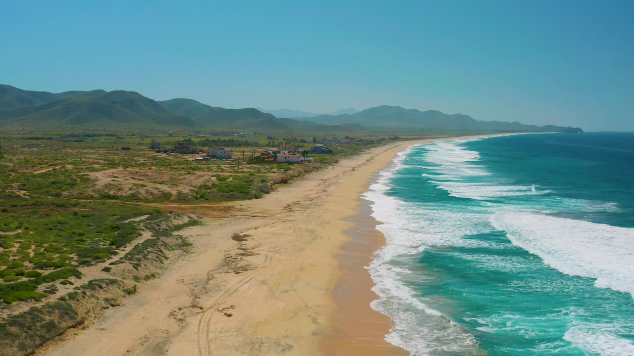 Wide aerial frame over Cerritos, Baja California Sur with repeating surf bands on the right, open sand below, green-brown vegetation inland, mountain ridges, and no visible people.