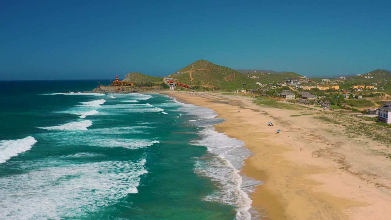 Opening aerial frame over Cerritos, Baja California Sur, with teal ocean and white surf on the left, wide tan beach on the right, coastal buildings and hills in the distance, and no visible people.