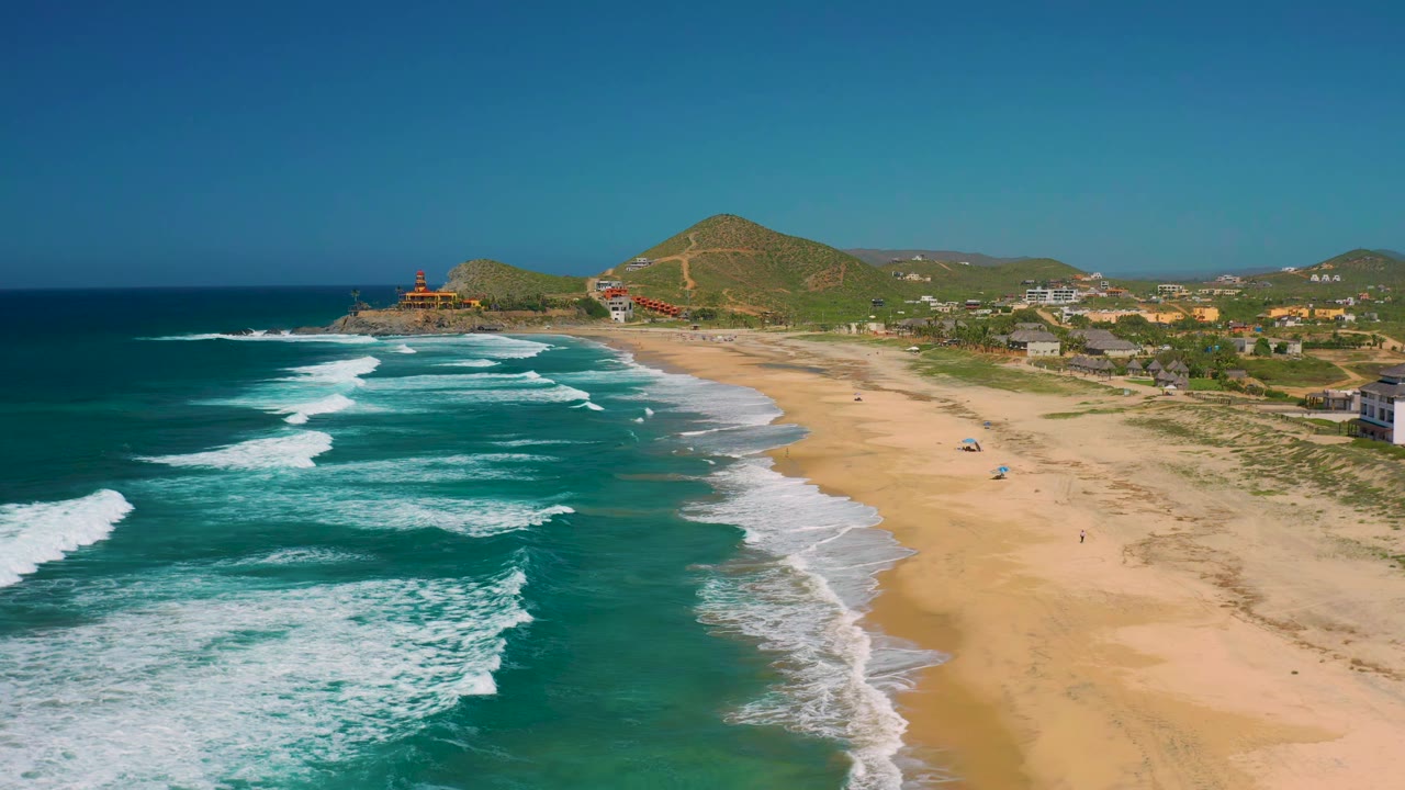 Drone view along a sandy coast with surf bands and hills beyond.