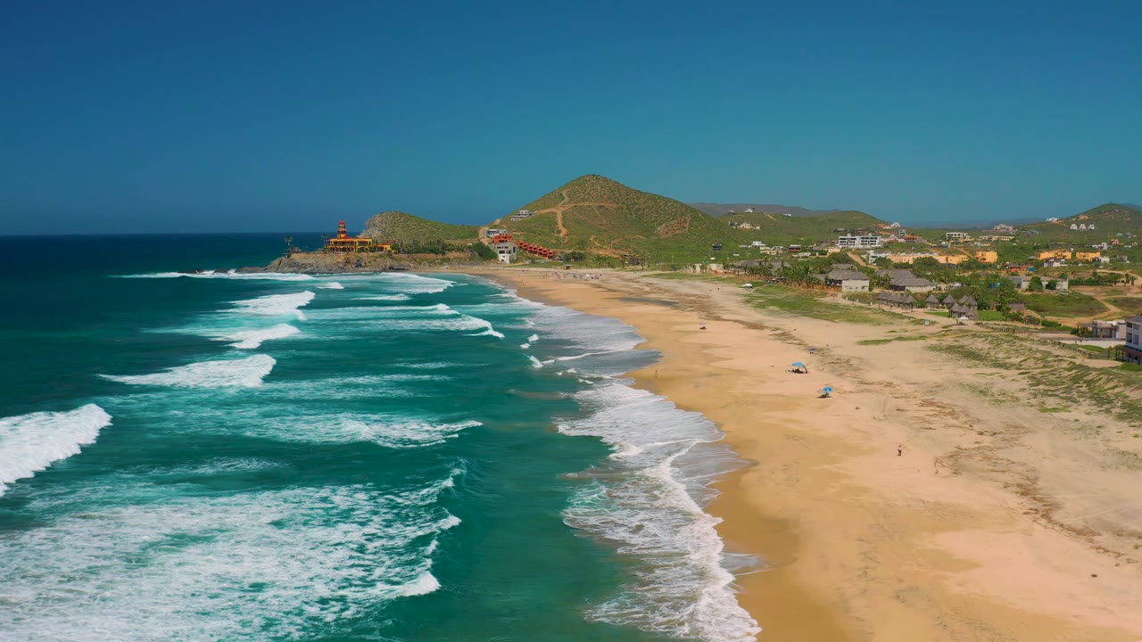 Cerritos coastline from an elevated drone angle, with turquoise water and foamy breakers in the foreground, open sand beside the waterline, hillside structures in the background, and no visible people.