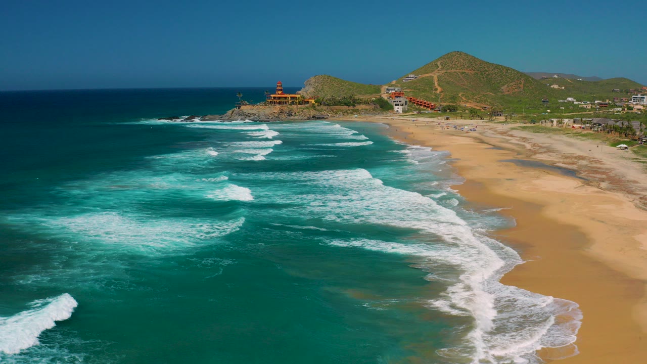 Aerial frame over Cerritos, Baja California Sur with turquoise water in the foreground, white surf bands reaching a wide tan beach, low green hills behind, and tiny beachgoers visible on the sand.