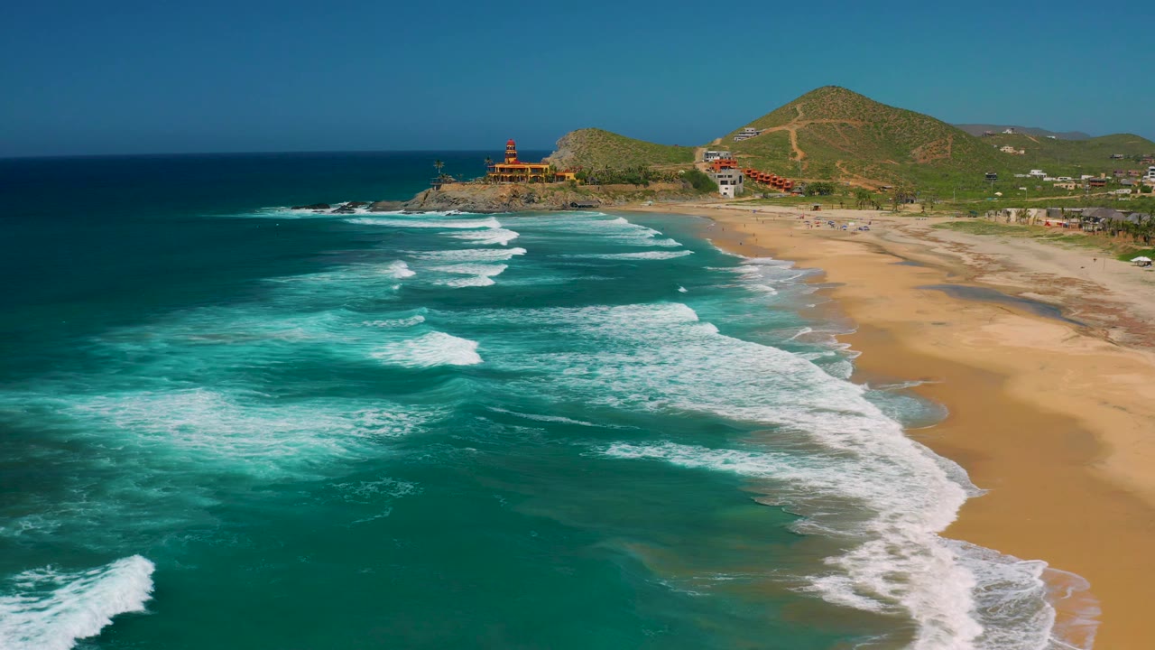 Wide aerial frame over Cerritos, Baja California Sur where white surf curves through the center, shallow turquoise water fills the left side, tan beach fills the right, and hillside paths and buildings appear in the background.