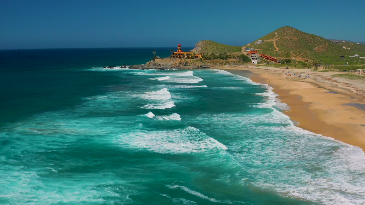 Aerial frame over Cerritos, Baja California Sur, with turquoise surf in the foreground, golden beach on the right, shoreline buildings near the point, green hills behind, and no visible people.