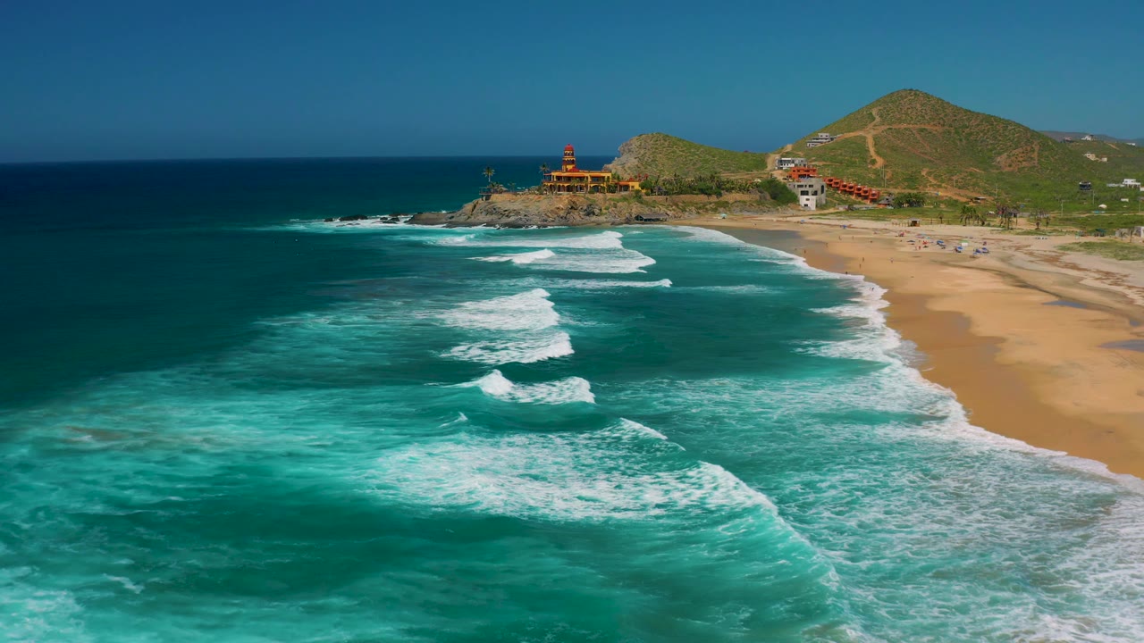 Cerritos coastline from above, with broad turquoise water across the frame, curved white surf approaching the beach, low green hills in back, shoreline structures near the point, and no visible people.