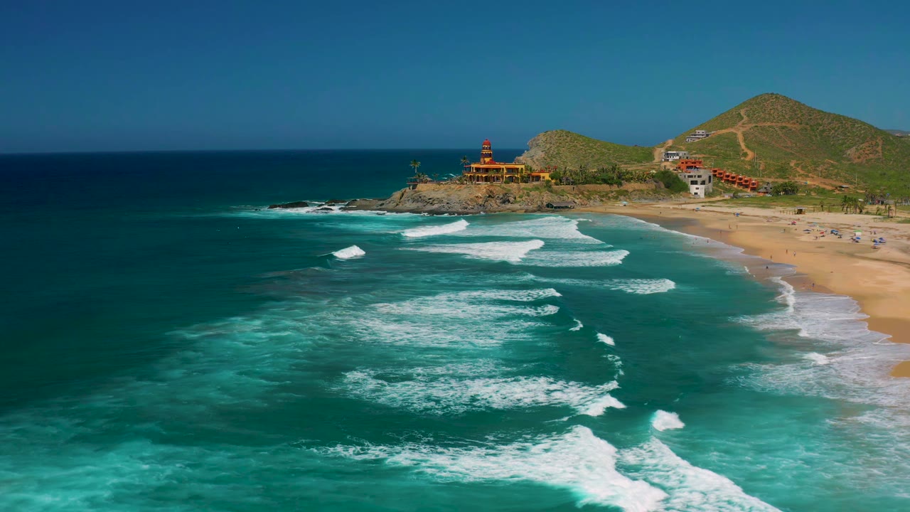 Aerial view in Cerritos, Baja California Sur with the surf bands shifted closer to shore, the rocky point and hillside buildings in the background, and no visible people.