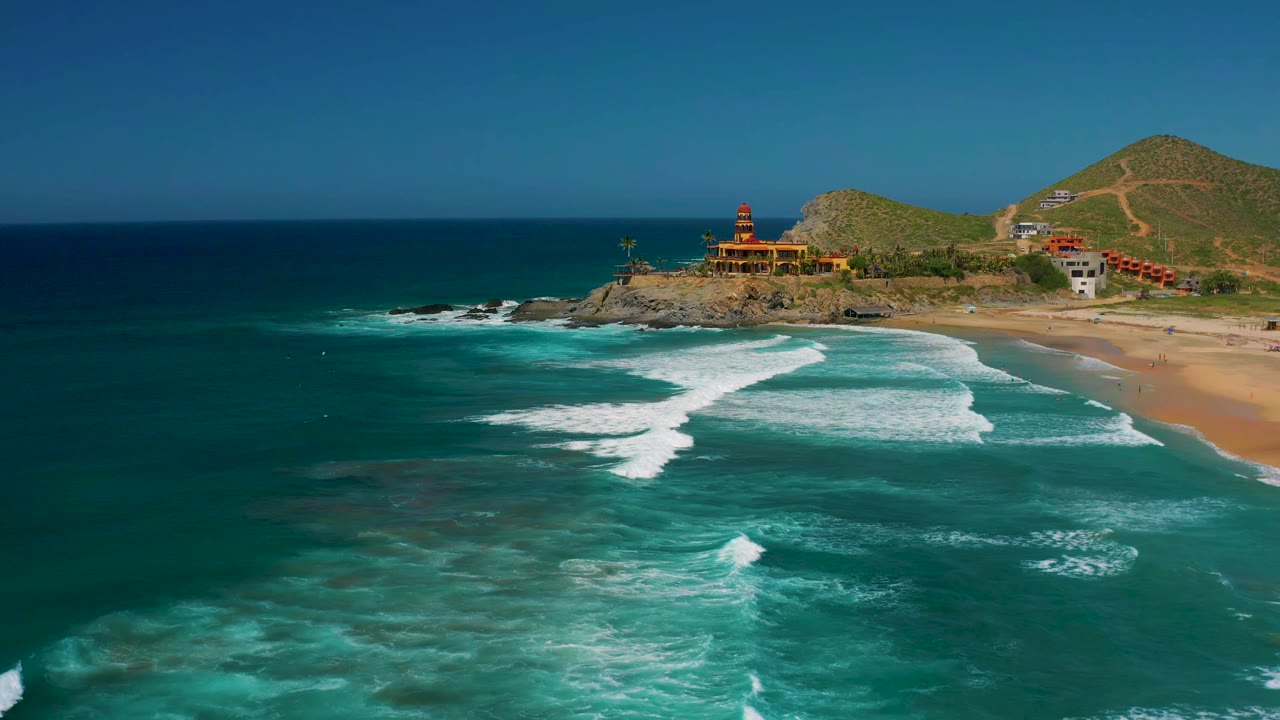 Drone view of Cerritos coastline with larger white surf bands in the center, dark blue offshore water, a sandy beach at right, hillside buildings, and no visible people.