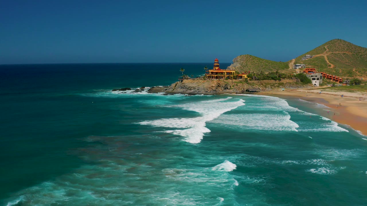 Aerial frame over Cerritos showing a rocky point with low buildings, a mountain ridge behind it, white surf moving across teal water, and no visible people.
