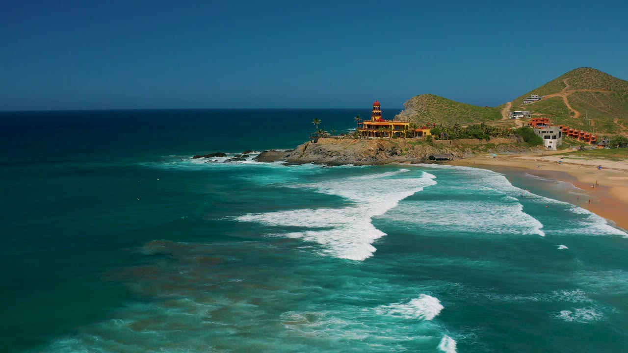 Cerritos aerial view with layered surf crossing the lower frame, sandy beach at right, a rocky shoreline with buildings above it, dry hills, and no visible people.