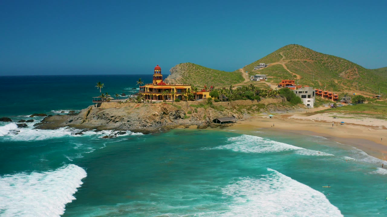Wide aerial frame in Cerritos, Baja California Sur showing a yellow and red building on a rocky point, turquoise water in the foreground, sandy beach to the right, green hills behind, and no visible people.