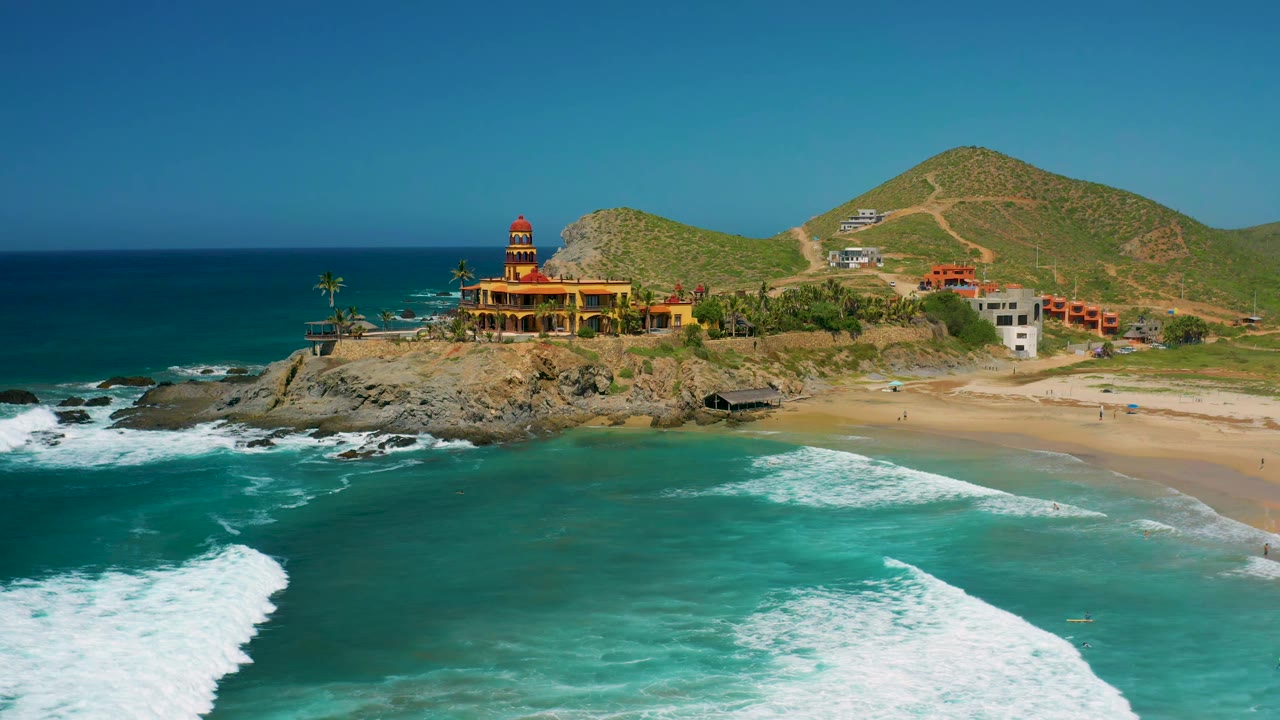 Aerial view in Cerritos, Baja California Sur with the coastal building centered left, white surf rolling across turquoise water, a sandy beach edge on the right, hillside roads in back, and no visible people.