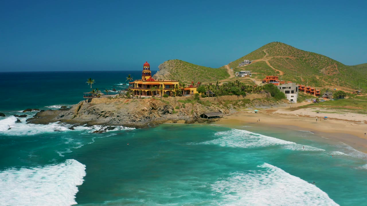 Cerritos, Baja California Sur aerial frame showing the beachfront building with palms beside it, foamy waves in the foreground, a pale sand beach to the right, green hills beyond, and no visible people.