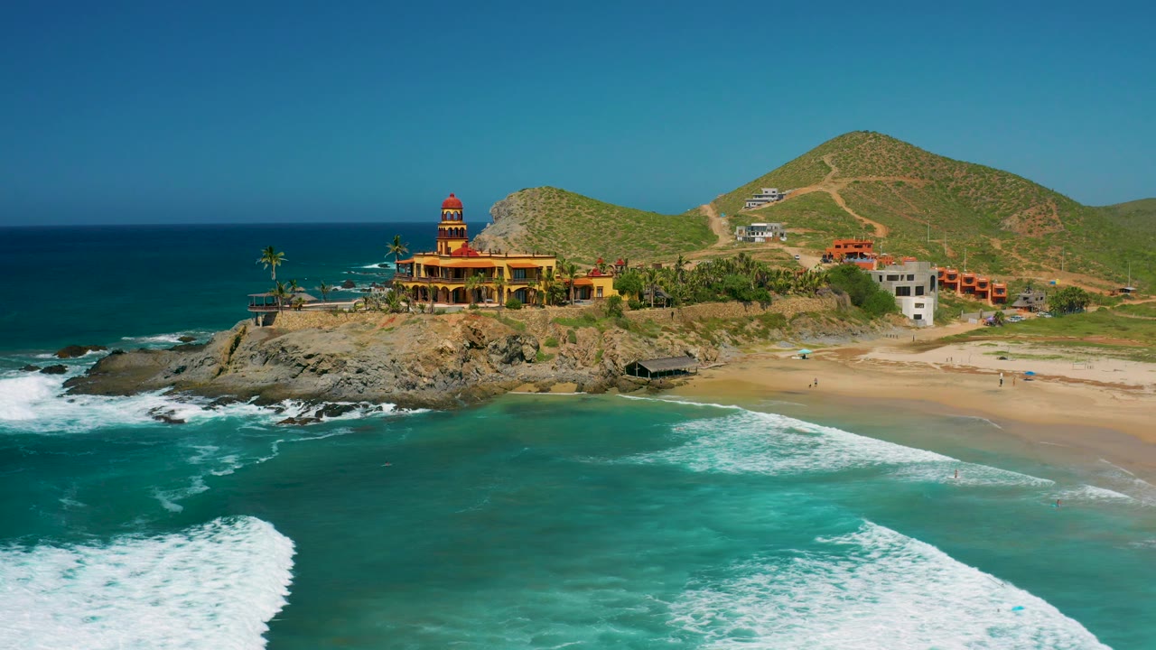 Wide coastal aerial with building, beach, surf, and hills