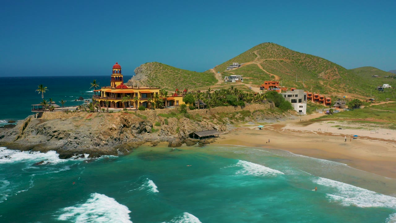 Opening frame over Cerritos, Baja California Sur shows turquoise water, white surf, a sandy beach, palms, a yellow-orange coastal building, hillside roads, and no visible people.