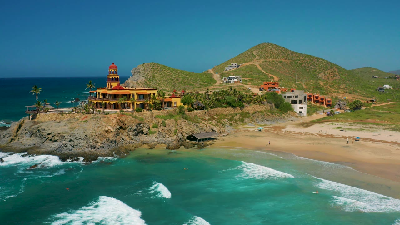 At 0:02, the Cerritos aerial view shows stronger white surf in the lower frame, the sandy beach to the right, the coastal building near the rocks, hills behind it, and no visible people.