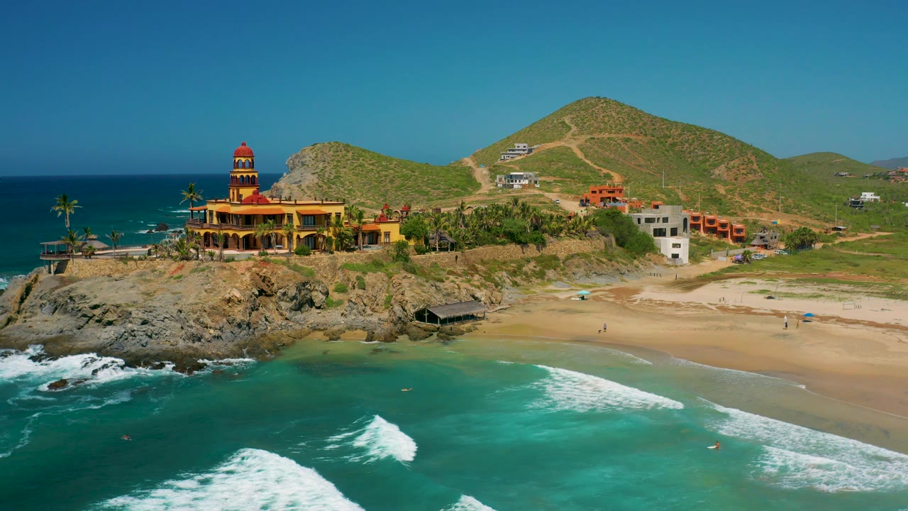 At 0:03 over Cerritos, Baja California Sur, surf fills more of the lower foreground while the beach, palms, yellow-orange building, hillside tracks, and no visible people remain in view.