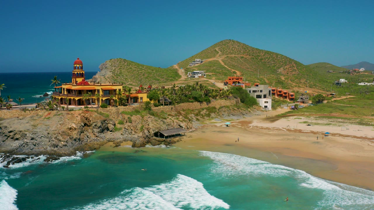 Wide aerial frame in Cerritos, Baja California Sur with turquoise surf in front, a sandy beach, rocky point, yellow mansion with red domed tower, hillside homes, and no discernible people.