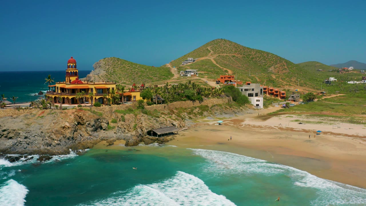Cerritos, Baja California Sur aerial frame with blue-green water and white surf below, a long sandy beach, rocky outcrop, large yellow mansion, hillside tracks, sparse buildings, and no discernible people.