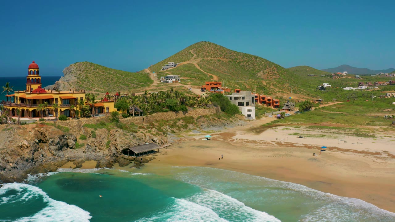 Wide aerial frame over Cerritos, Baja California Sur with foamy surf below, open sand, palm trees near a yellow building, hillside roads, scattered homes, and no clearly visible people.