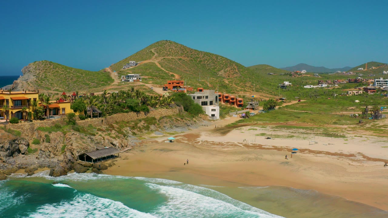 Aerial view of Cerritos, Baja California Sur with turquoise surf in the foreground, a sandy beach, rocky shore, scattered homes, green hills, and tiny distant people on the sand.