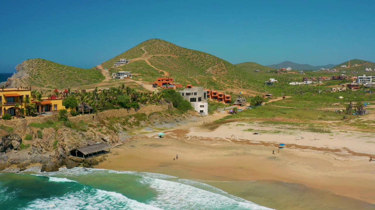 Aerial view in Cerritos, Baja California Sur with white foam along the lower edge, open sand, rocky coastline, beachfront houses, palms, and low green hills under clear daylight.
