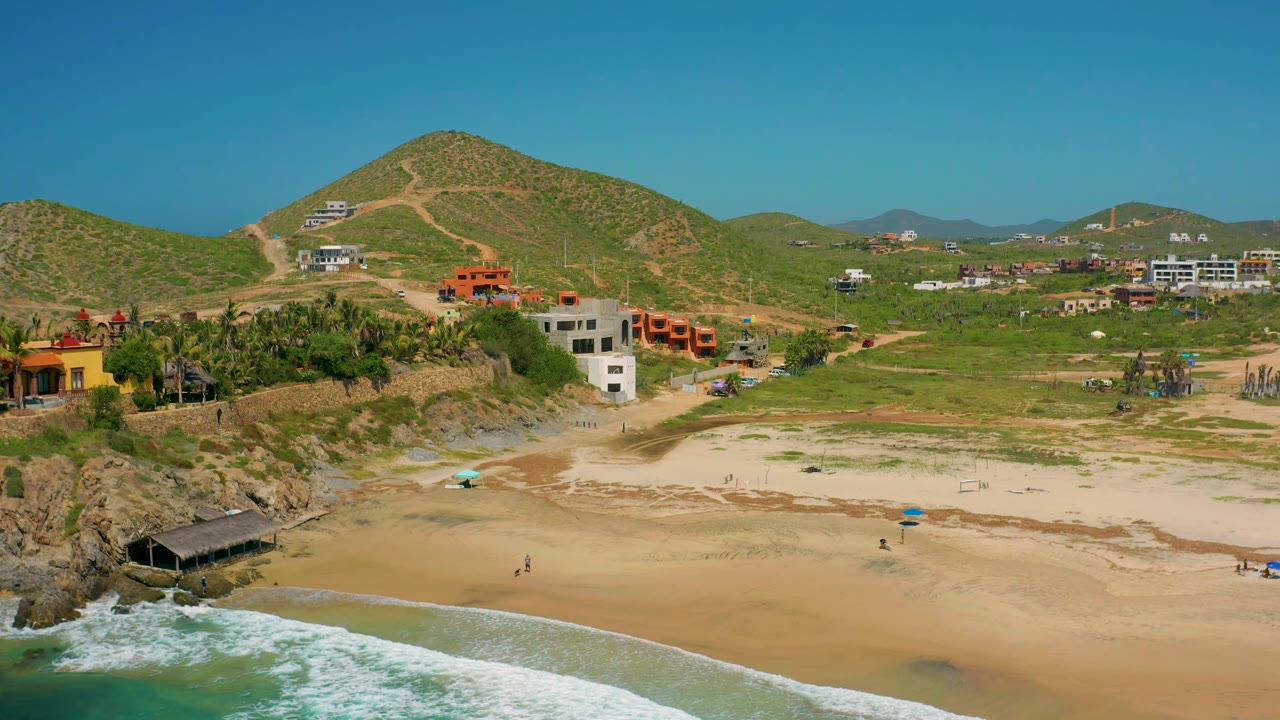 Aerial view over Cerritos beach showing white surf at the lower edge, pale sand, a rocky point, palms, low buildings, green hills, and no visible people.