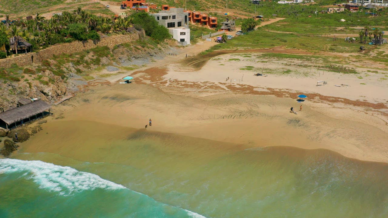 Aerial view over Cerritos, Baja California Sur showing green-blue surf at the lower edge, wet sand, rocky shoreline, beach umbrellas, low buildings, and small distant people on the beach.
