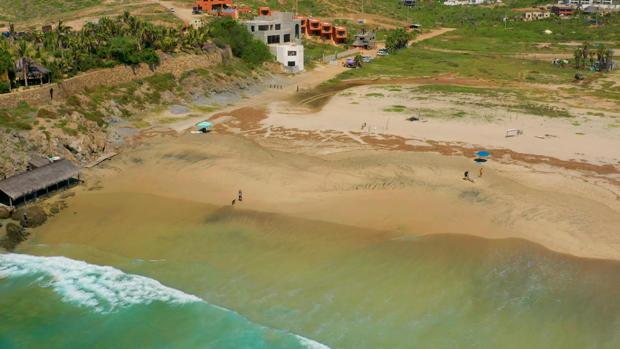 High aerial frame over Cerritos with a curling band of white surf in the lower left, tan wet sand across the center, rocks and a dark palapa at left, and small distant people near the waterline.