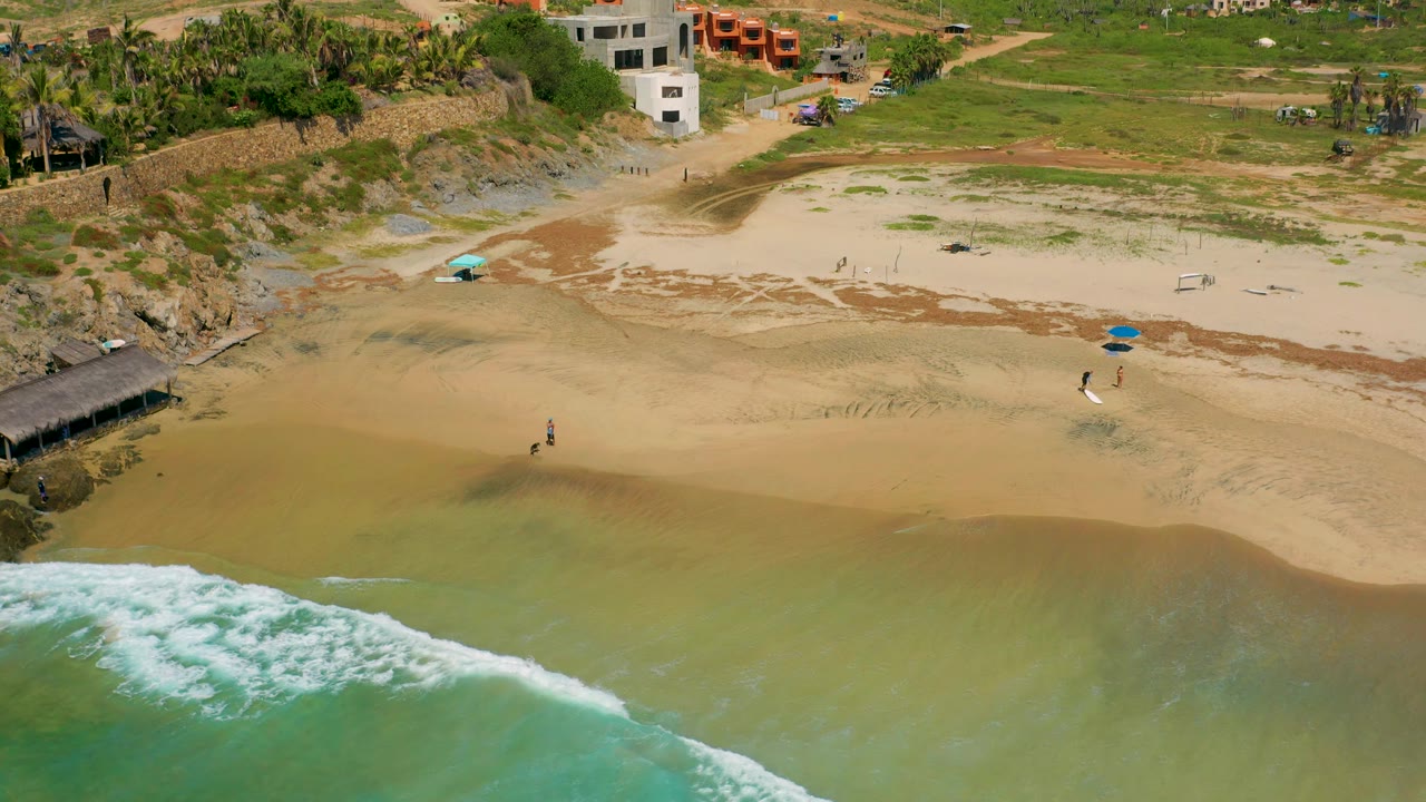 Aerial view of Cerritos shoreline with foamy surf entering from the lower left, broad wet sand, scattered umbrellas, a rocky outcrop, hillside vegetation, and low buildings behind the beach.
