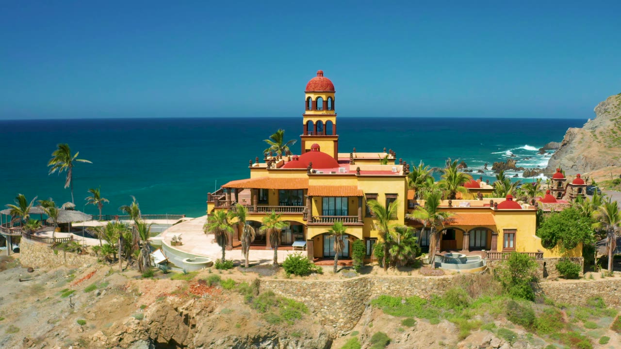 A steady aerial frame shows the Cerritos coastal building centered, with terraced walls, palms, surf behind it, a rocky slope to the right, and no visible people.
