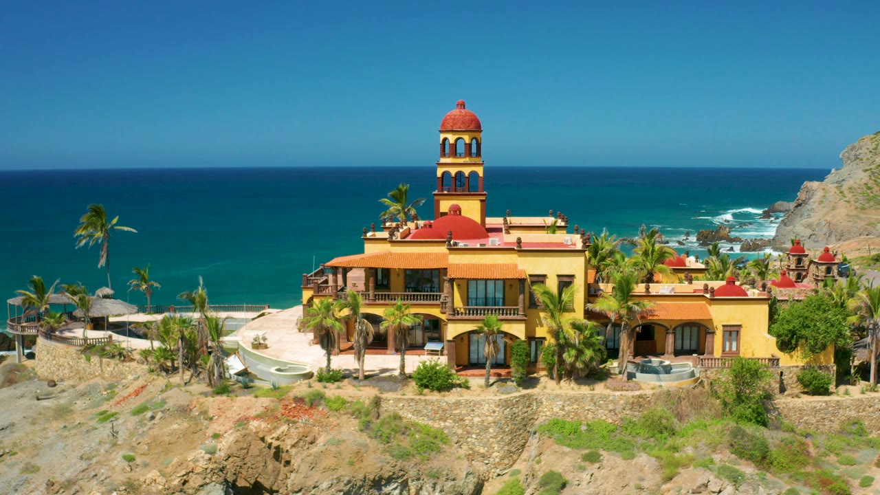 The Cerritos property remains framed from above, with the red tower roof near center, palms along the terrace, turquoise water behind, and no visible people.
