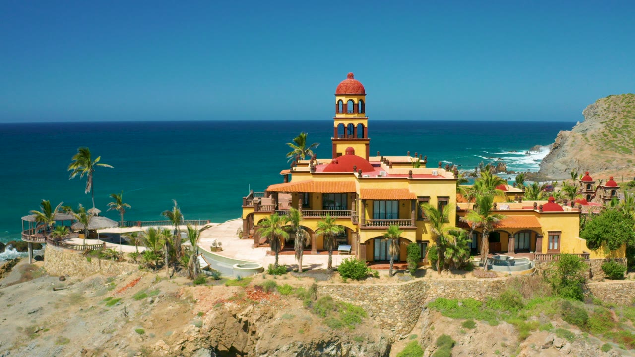 Cerritos, Baja California Sur coastal building with a terracotta tower, palms, rocky foreground, blue ocean, distant headland, and no visible people at the start of the clip.