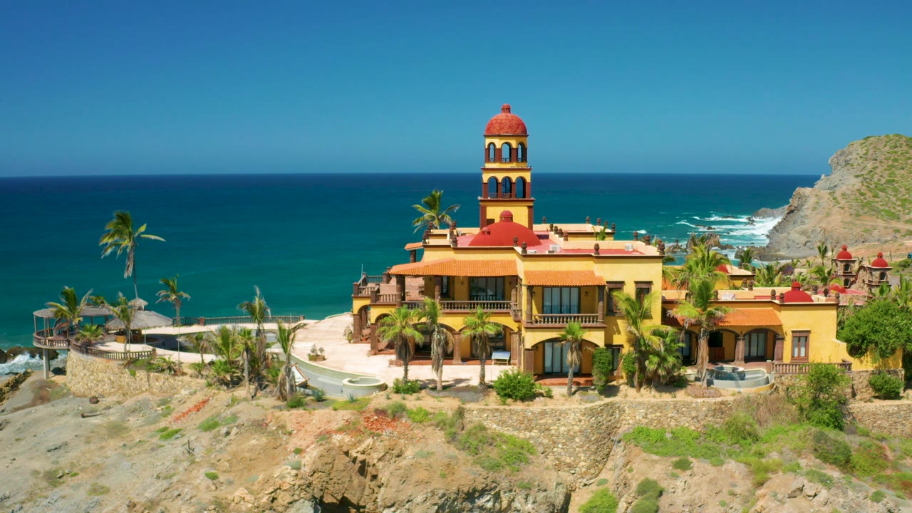 Cerritos, Baja California Sur frame showing the large yellow building, red roof sections, palms, open sea, right-side headland, and no visible people.