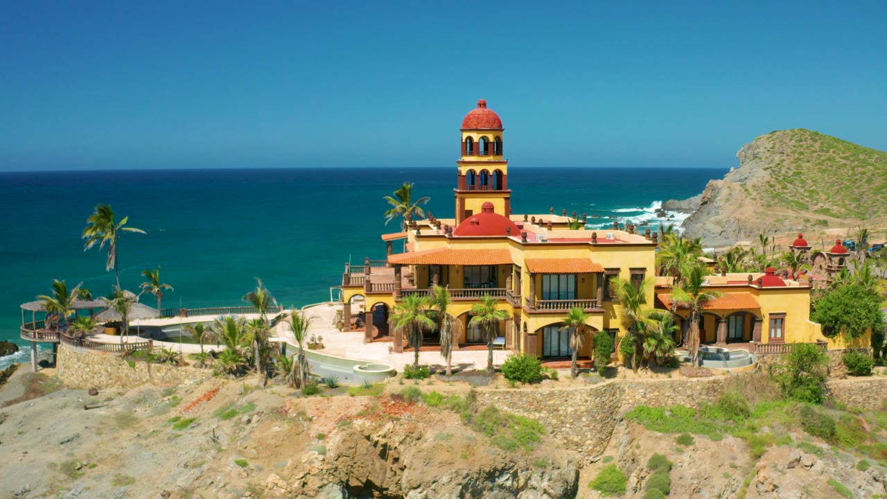 Wide aerial view in Cerritos, Baja California Sur of a yellow and terracotta house with a tower, palm trees, rocky sand, turquoise ocean, and no visible people.