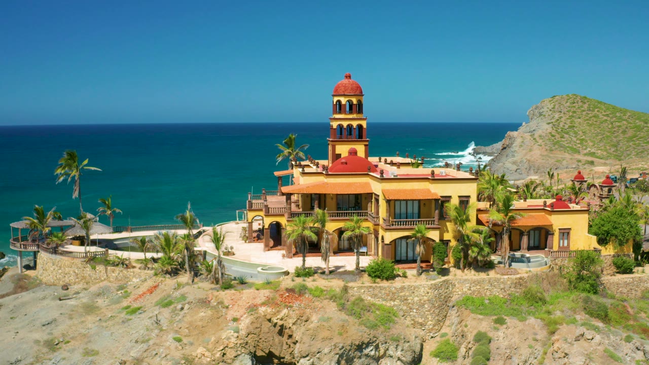 Aerial 4K frame from Cerritos showing the yellow house, red roof details, palms, surf line, rocky terrain, hillside, and no visible people.