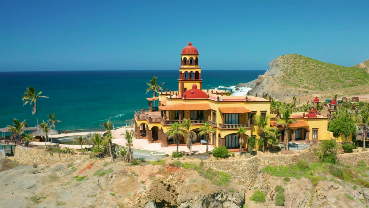 Elevated Cerritos frame with the red-roofed tower centered above the yellow building, palm trees at the beach edge, blue ocean behind, hillside to the right, and no visible people.