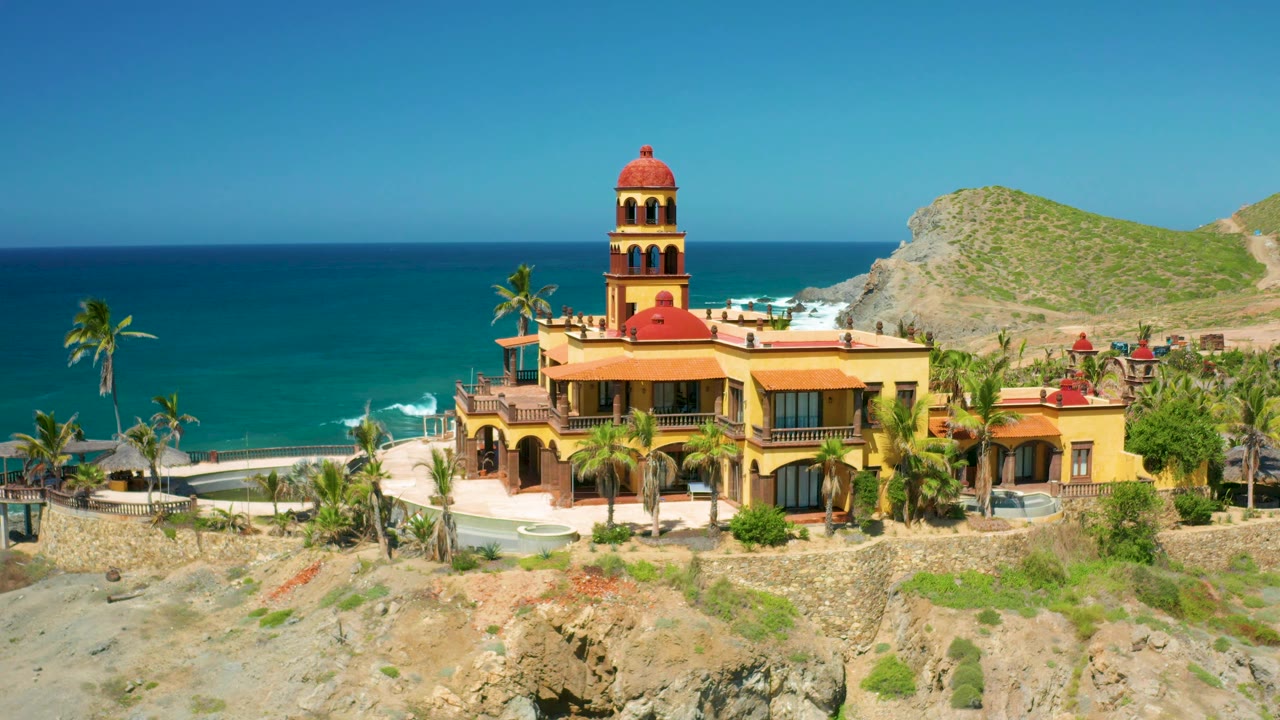 Wide view in Cerritos, Baja California Sur of the yellow coastal property, red tower dome, palms, beach surf, rocky slope in front, ocean horizon, and no visible people.