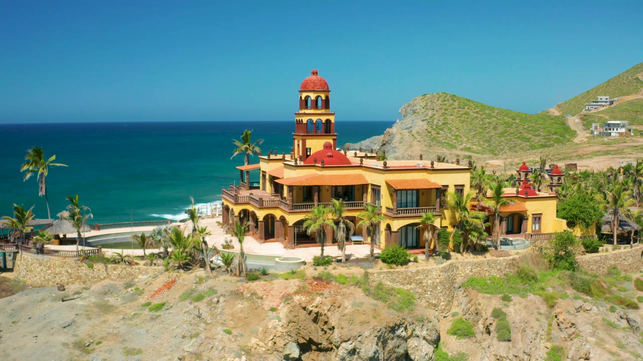 A decorative yellow building with red roof accents sits near the Cerritos shoreline, with palms below, a blue ocean band behind it, dry hills at right, and no visible people.