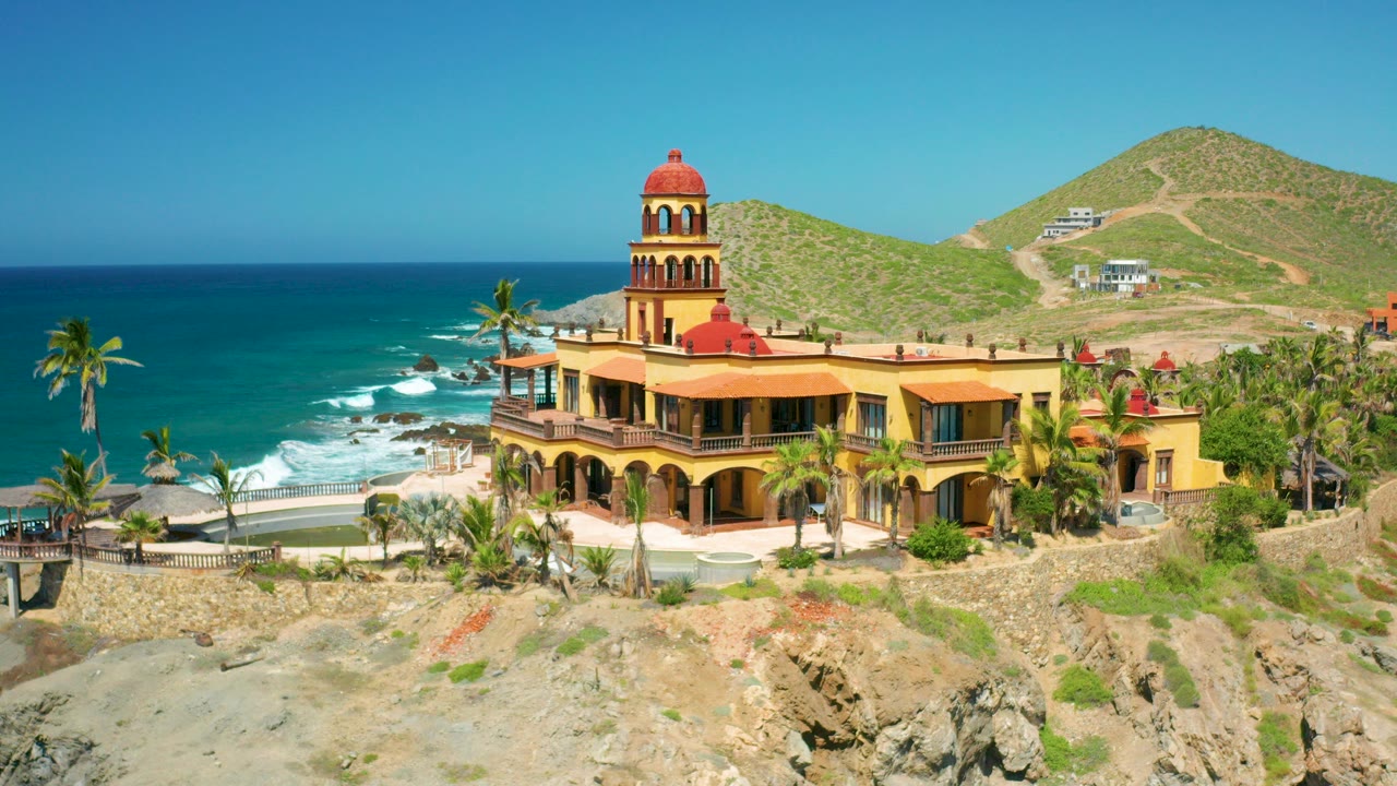 Opening frame of a yellow Cerritos, Baja California Sur coastal house with a terracotta tower, rocky foreground, blue ocean to the left, hills behind, and no visible people.