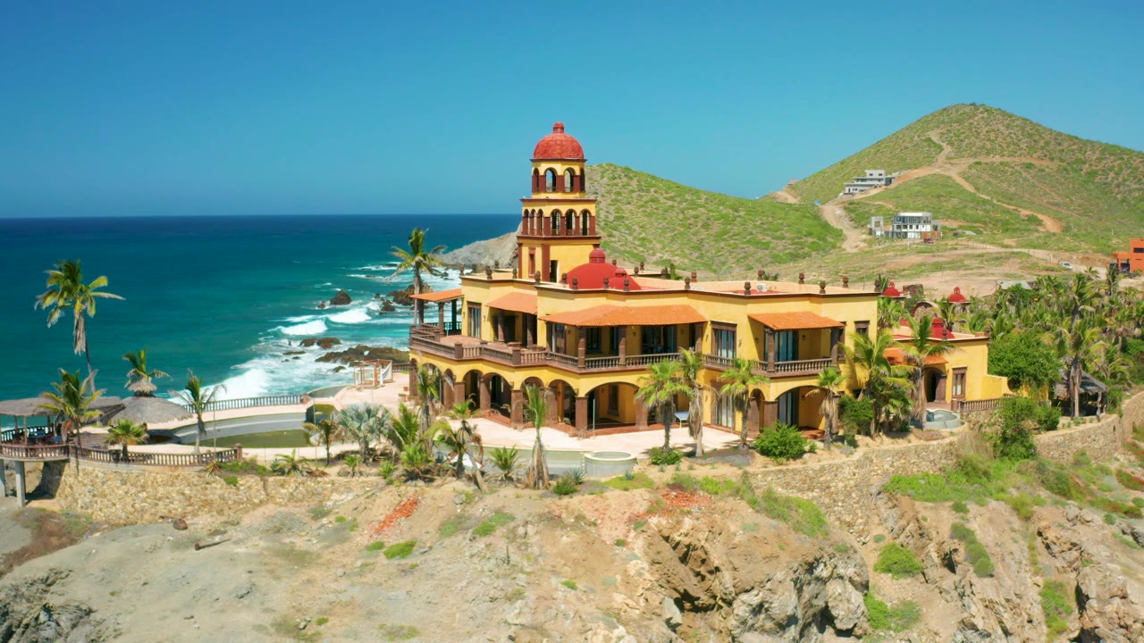 Third frame of the Cerritos house and tower, with the ocean horizon running behind the palms, white water along the shore, green hills in back, and no visible people.