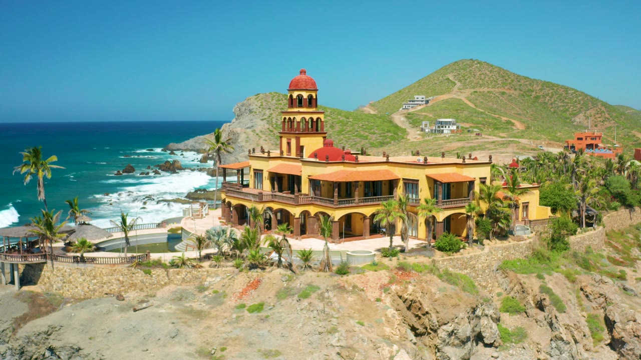 At 0:02, the aerial composition shows the red-domed tower above the yellow structure, palms around the property, ocean spray to the left, and Cerritos hills behind, with no visible people.