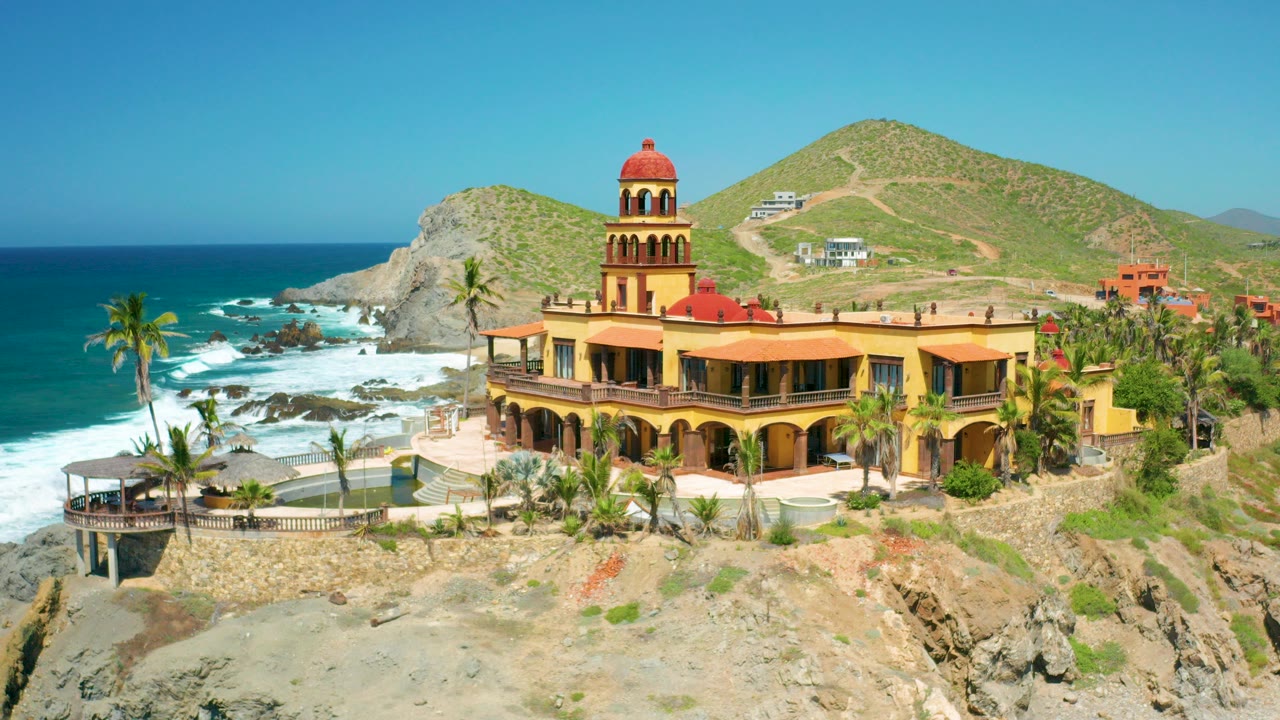 Opening aerial frame in Cerritos, Baja California Sur showing a yellow church-like building with a red dome on rocky cliffs, blue ocean to the left, palms below, and no visible people.
