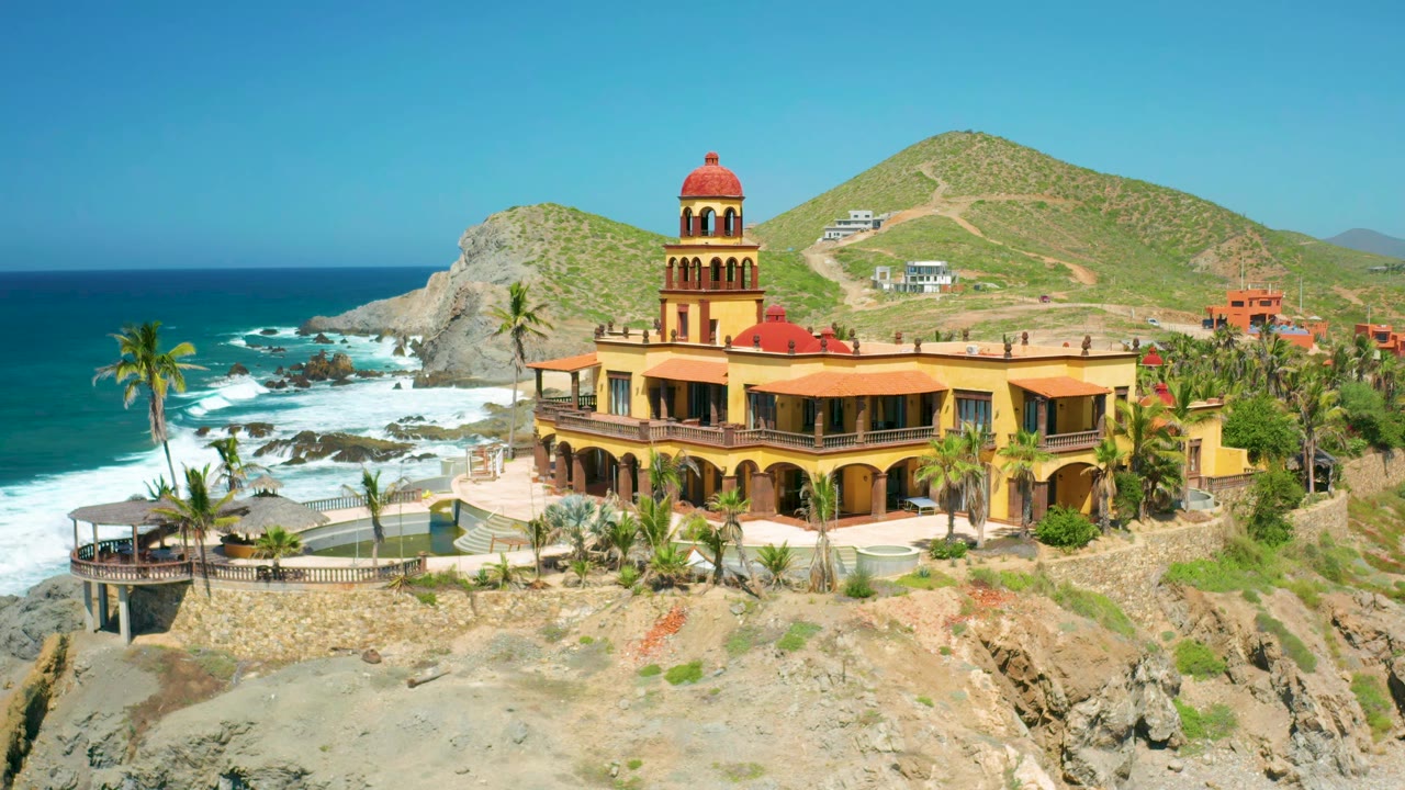 Aerial frame in Cerritos, Baja California Sur with the red-domed church centered over cliffside terraces, waves breaking on rocks at left, green hills behind, and no visible people.